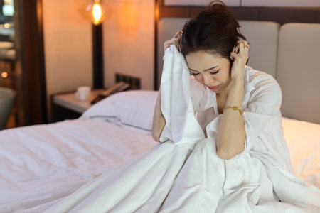 Young depressed asian woman sitting and holding head with hands while crying on bed in hotel bedroom with low light environment. Sad girl, unhappy and loneliness concept.の写真素材