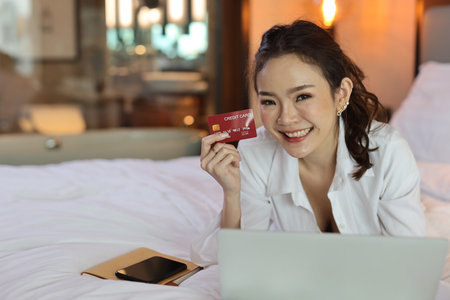 Happy asian woman in casual lying on bed and holding credit card while using computer in hotel for online shopping payment, beauty businesswoman with bedroom background. Lifestyle technology conceptの写真素材