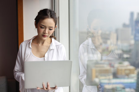 Portrait of confident and beautiful young asian businesswoman with happy emotion standing and using computer in modern hotel with city blurred background. Success business and lifestyle conceptの写真素材