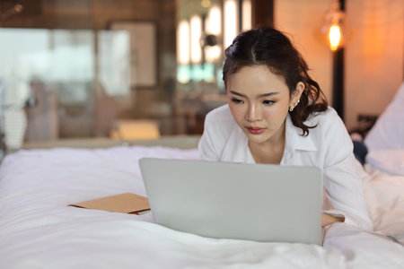 Active asian woman in casual lying on bed and working on computer with notebook in hotel, beauty businesswoman with bedroom background. Lifestyle technology conceptの写真素材