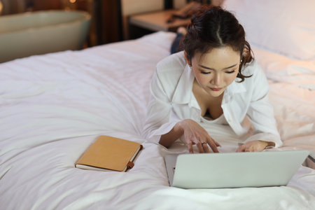 Active asian woman in casual lying on bed and working on computer with notebook in hotel, beauty businesswoman with bedroom background. Lifestyle technology conceptの写真素材
