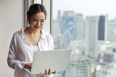 Portrait of confident and beautiful young asian businesswoman with happy emotion standing and using computer in modern hotel with city blurred background. Success business and lifestyle conceptの写真素材