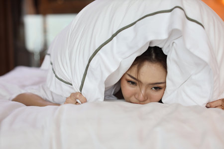 Young depressed asian woman lying on bed with pillow on head in bed at hotel bedroom with low light environment. Sad girl, unhappy and loneliness concept.の写真素材