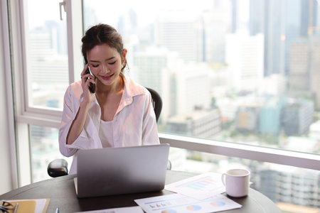 Portrait of confident and beautiful young asian businesswoman with happy face sitting and using computer and mobile smartphone in hotel with city background. Success business and lifestyle conceptの写真素材