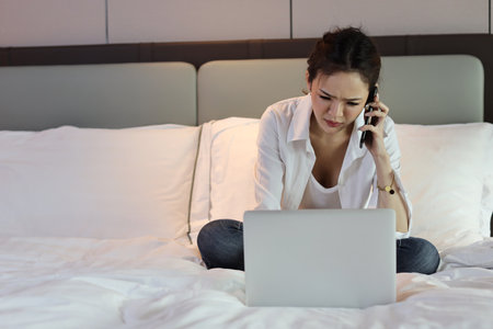 Active and unhappy asian woman in casual sitting on bed and working smart mobile phone and computer in hotel, beauty businesswoman with bedroom background. Lifestyle technology conceptの写真素材