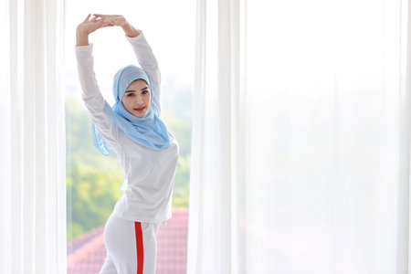 Beautiful asian woman wearing white muslim sleepwear, stretching her arms after getting up in the morning at sunrise. Cute young girl with blue hijab standing and relaxing with happy and smiling face.の写真素材
