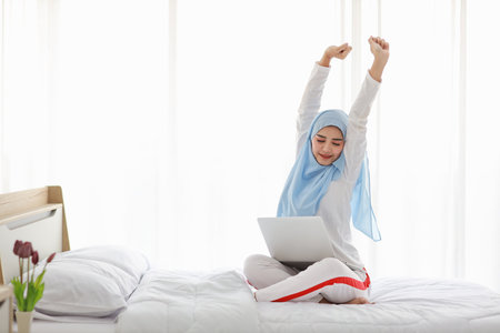 Young asian arab woman using laptop sitting on bed in bedroom. Beautiful girl in muslim sleepwear with blue hijab stretching her arms after working before go to bed. Lifestyle and technology conceptの写真素材