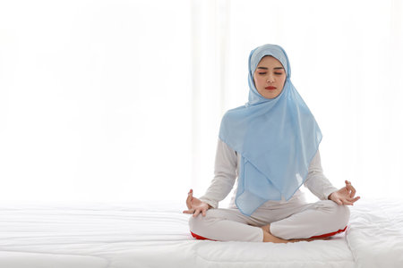 Young asian woman sitting on bed and enjoying meditation. Beautiful girl in muslim sleepwear with blue hijab practices yoga in bedroom with peace and calm. Healthy and lifestyle conceptの写真素材