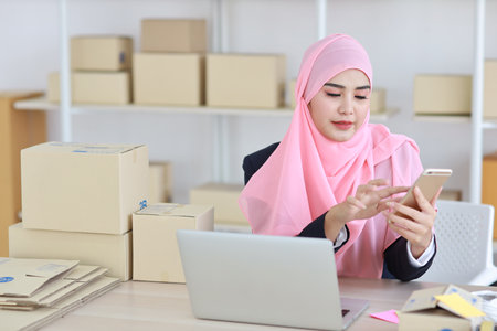 Religious asian woman in muslim dress sitting and using smart mobile phone and talking to customer with happy face and business package SME box delivery background.の写真素材