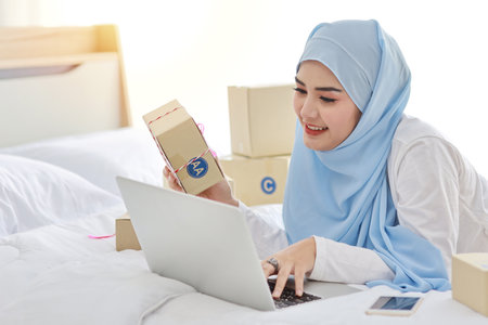 Beautiful and young asian woman in muslim sleepwear with attractive look, lies on bed with computer and online package box delivery. Startup small business SME freelance girl working with mobile phoneの写真素材