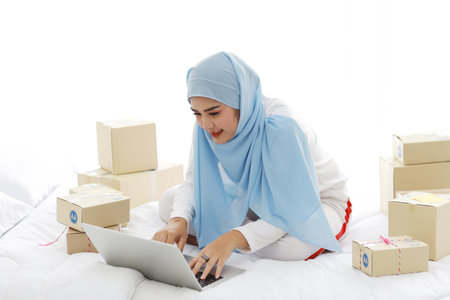 Beautiful and young asian woman in muslim sleepwear with attractive look, sitting on bed with computer and online package box delivery. Startup small business SME freelance girl work with mobile phoneの写真素材