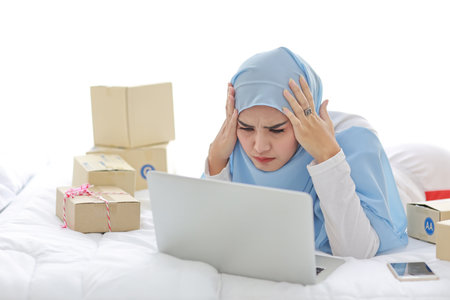 Beautiful and young asian woman in muslim sleepwear with headache emotion lies on bed with computer, mobile phone and online package box delivery. Smart girl with hijab receive bad news and tiredの写真素材