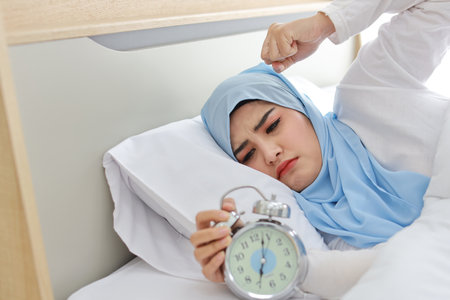 Oversleeping asian woman white muslim wearing sleepwear lying on bed, missing ring of alarm clock waking up. Portrait young girl trying to stop ringing in the morningの写真素材
