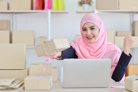 Active asian woman in blue muslim suit sitting and working with computer and showing online package box delivery with happy smiling face. Startup small business freelance working girl look at product.の写真素材