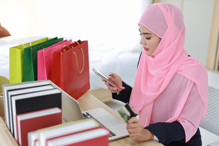 Active smart asian woman in blue muslim suit and pink shaft sitting and holding credit card while using mobile phone for payment or online purchasing. Startup small business girl work at home.の写真素材
