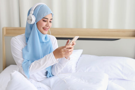 Portrait of beautiful asian female in muslim sleepwear watching online story on mobile phone, lites on bed and connected with wireless internet. Young cute girl with hijab listen music from smartphoneの写真素材