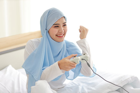 VDO game console station concept. active asian woman wearing white muslim sleepwear sitting on bed, holding joystick and playing exciting game. Cute girl looked excited with game controller consoleの写真素材