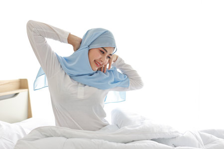Beautiful asian muslim woman wearing white sleepwear sitting on bed, stretching her arms after getting up in the morning at sunrise. Cute young girl with hijab wake up and relaxing, closing her eyesの写真素材