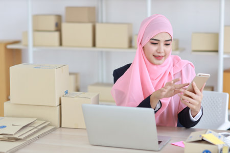 Religious asian woman in muslim dress sitting and using smart mobile phone and talking to customer with happy face and business package SME box delivery background.の写真素材