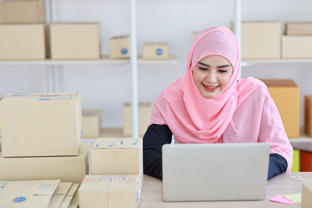 Active asian woman in blue muslim suit sitting and working with computer and online package box delivery. Startup small business freelance working girl look at camera with smilingの写真素材