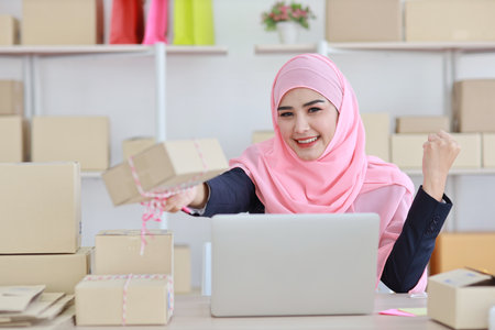 Active asian woman in blue muslim suit sitting and working with computer and showing online package box delivery with happy smiling face. Startup small business freelance working girl look at product.の写真素材