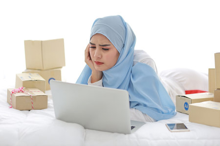 Beautiful and young asian woman in muslim sleepwear with headache emotion lies on bed with computer, mobile phone and online package box delivery. Smart girl with hijab receive bad news and tiredの写真素材