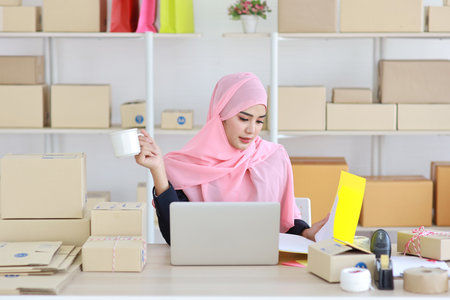 Active smiling asian woman in blue muslim suit sitting and working with online package box delivery. Startup small business SME freelance girl working on computer and mobile phone with happy face.の写真素材