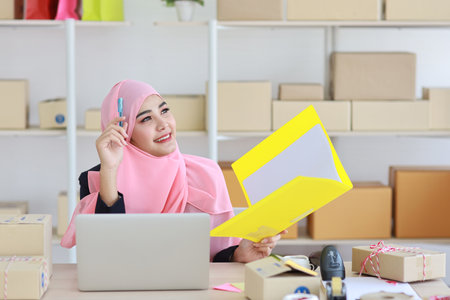Active asian woman wearing muslim suit sitting and holding folder with computer and online package box delivery. Startup small business freelance concept.の写真素材