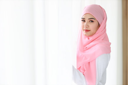 Beautiful asian woman wearing white muslim sleepwear, standing at window after getting up in the morning at sunrise. Cute young girl with pink hijab relaxing while looking at cameraの写真素材
