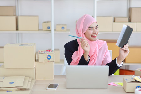 Active asian woman in muslim suit sitting and working with computer, mobile smartphone and online package box delivery. Startup small business freelance working girl checking customer order from bookの写真素材