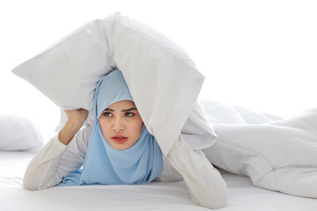 Oversleeping asian woman in muslim sleepwear lying on bed and missing ring of alarm clock waking up. Young girl trying to sleep by putting pillow over her head in bedroom with unhappy emotionの写真素材