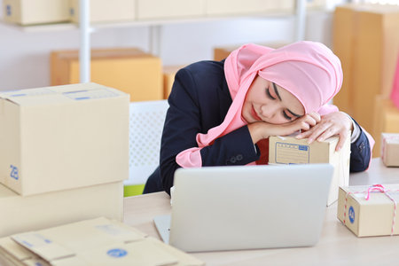 Religious asian woman in blue muslim suit and pink shaft frustrated laid her head down on the table, sit and work on wooden table with computer and package box delivery. Young girl sleep while workingの写真素材