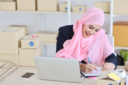 Active asian woman in muslim suit sitting and working with computer, mobile smartphone and online package box delivery. Startup small business freelance working girl checking customer order from bookの写真素材