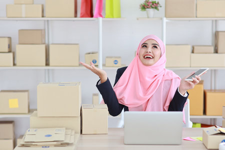 Active asian woman with muslim suit sitting and working with computer, mobile smartphone and online package box delivery. Startup small business freelance working girl showing something on her hand.の写真素材