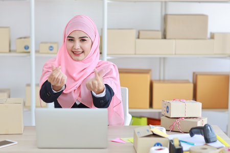 Active asian woman in blue muslim suit sitting and working with computer and online package box delivery. Startup small business freelance working girl showing love symbol with exciting emotion.の写真素材