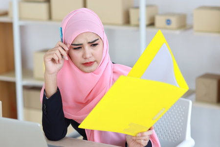 Active asian woman wearing muslim suit sitting and holding folder with seriously look and computer and online package box delivery on table. Startup small business freelance concept.の写真素材