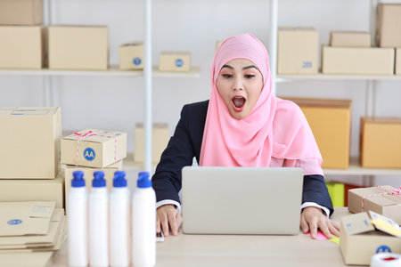 Active asian woman in blue muslim suit sitting and working with computer and online package box delivery. Startup small business freelance working girl look at camera with exciting emotion.の写真素材