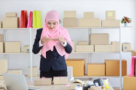 eligious asian woman in blue muslim suit standing and taking picture of package box deliver from mobile phone. Startup small business SME freelance girl work at home with happy smiling faceの写真素材