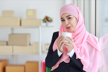 Religious asian woman in muslim blue suit drinking coffee, smiling and pink scarf on head with package box delivery background. Startup small business SME freelance girl work at home with happy faceの写真素材