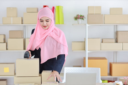 Active asian woman wearing muslim suit standing and using scanner with package box delivery. Startup small business SME freelance girl work at home concept.の写真素材