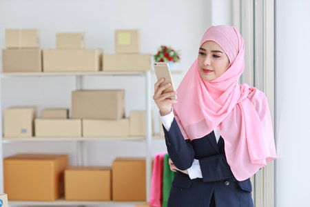 Religious asian woman in blue muslim suit and pink shaft on head standing and using mobile phone with confidence. Business woman stand with package SME box delivery background. Work at home conceptの写真素材