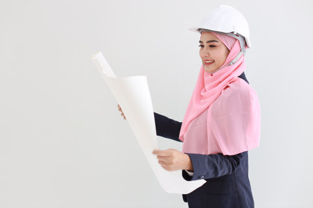 Smart young asian woman wearing blue muslim suit smiling confident holding blueprint in studio. Isolated white background portrait with beautiful face girl with pink hijab. Advertisement concept.の写真素材