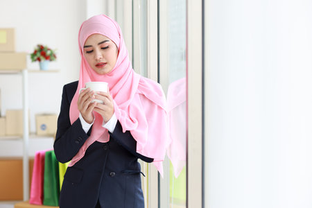 Religious asian woman in muslim blue suit drinking coffee, smiling and pink scarf on head with package box delivery background. Startup small business SME freelance girl work at home with happy faceの写真素材