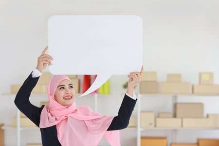 Active asian woman in muslim blue suit standing and holding white speech bubble with online package box delivery background. Startup small business SME freelance girl work at home with happy face.の写真素材