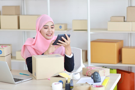 Active smiling asian woman in blue muslim suit sitting and working with online package box delivery. Startup small business SME freelance girl working on computer and mobile phone with happy face.の写真素材