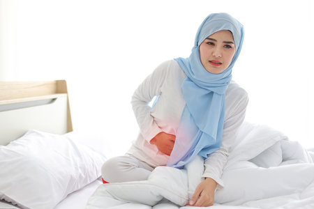 Menstrual pain or stomachache young asian muslim woman sitting on bed. Beautiful girl hold stomach and feeling painful with unhappy face. Healthy conceptの写真素材