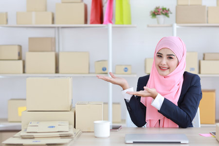 Active asian woman in blue muslim suit sitting and working with computer and online package box delivery. Startup small business freelance working girl showing something with exciting emotion.の写真素材
