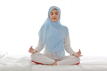 Close up young asian arab woman sitting on bed and enjoying meditation. Beautiful girl in muslim sleepwear with blue hijab practices yoga in bedroom with peace and calm. Healthy and lifestyle conceptの写真素材