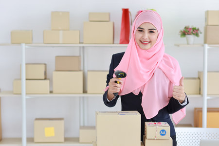 Active asian woman wearing muslim suit standing and using scanner and showing thumb up with package box delivery. Startup small business SME freelance girl work at home concept.の写真素材