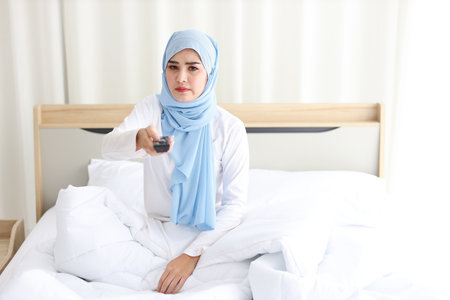 TV and happiness concept. Beautiful asian woman in white muslim sleepwear sitting on bed, holding television remote and raise hand, shout loudly and celebrate something with glad happy smiling face.の写真素材